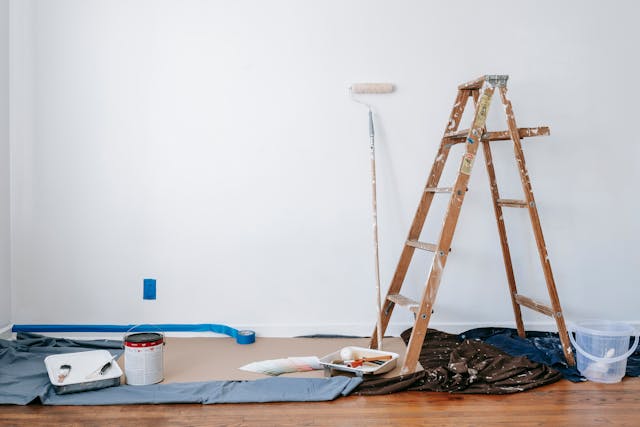 Cuarto de hogar con un muro al fondo pintado con pintura vinílica color blanco y en el piso se observa equipo y utensilios de trabajo de un oficial pintor.